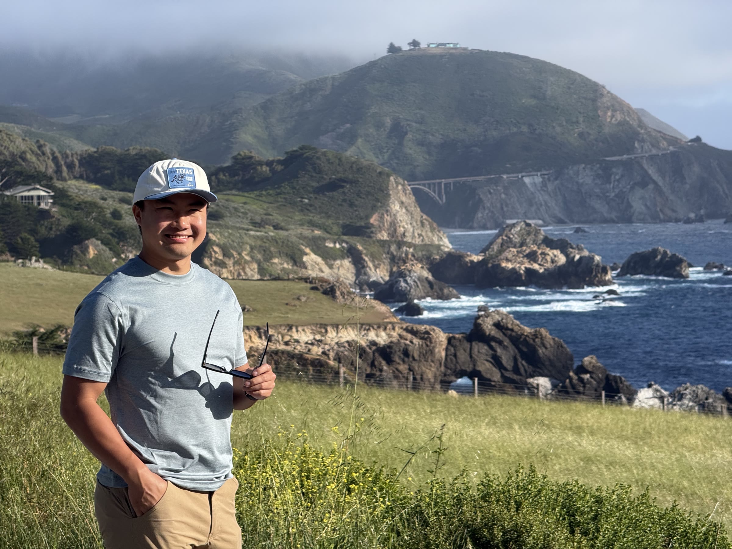 Tony Kam smiling on a grassy overlook above Bixby Creek Bridge and the California coast
